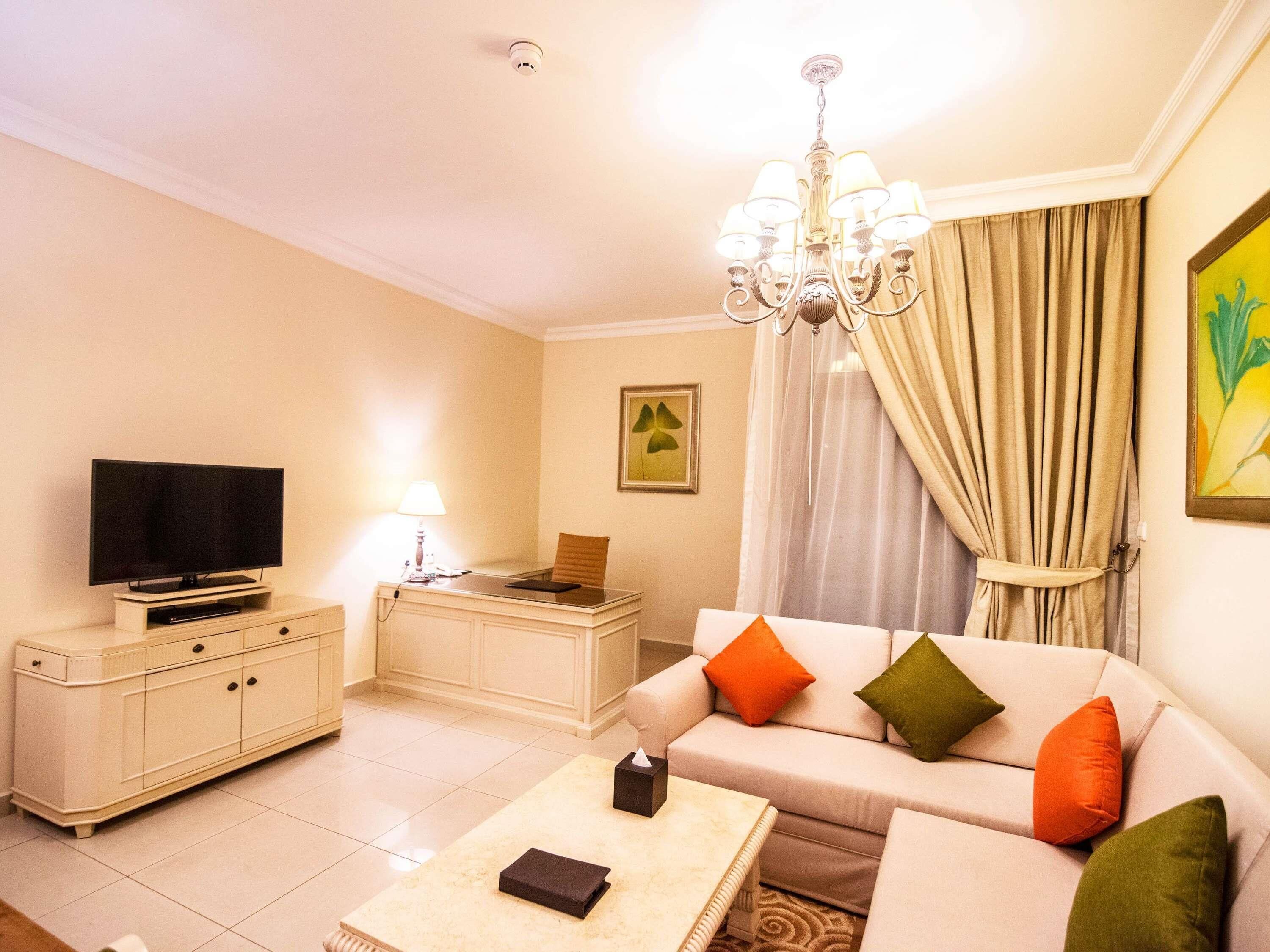 A living room with a television, a desk, a sofa with cushions, a chandelier, and a painting on the wall.