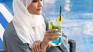 Une personne assise au bord d’une piscine, un verre à la main, portant un foulard blanc et une robe grise.