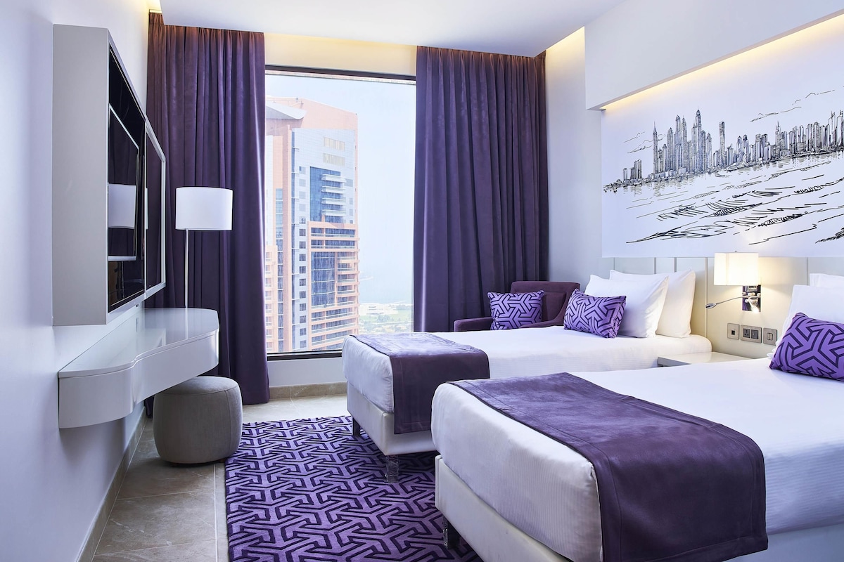 Quarto de hotel moderno com duas camas, uma janela grande com cortinas, um mural com vista para a cidade e uma televisão de tela plana.