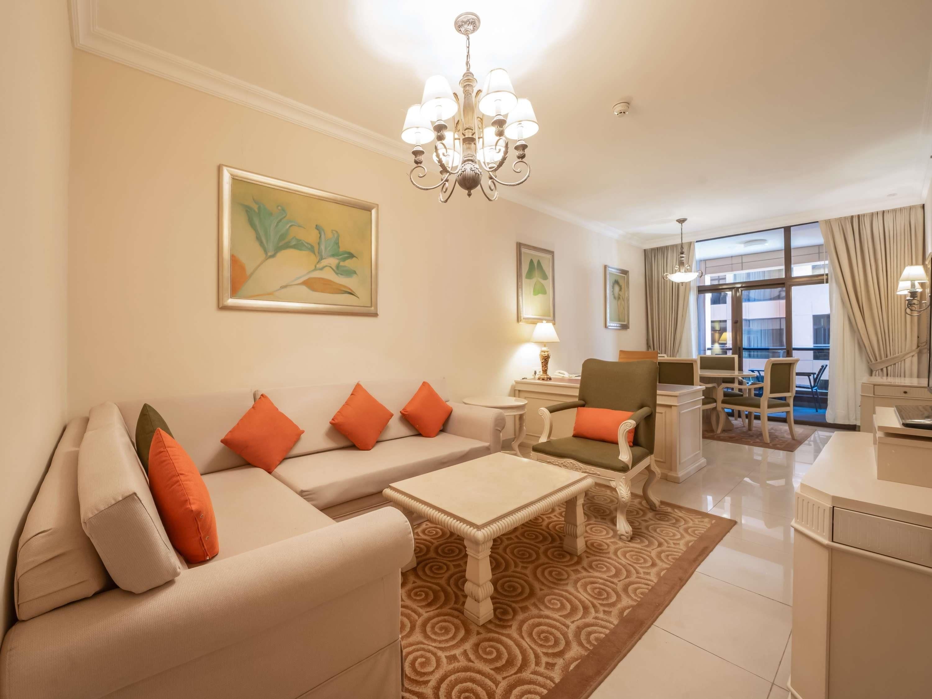 A hotel room with a sofa, armchairs, a coffee table, a dining area with chairs, and artwork on the walls.