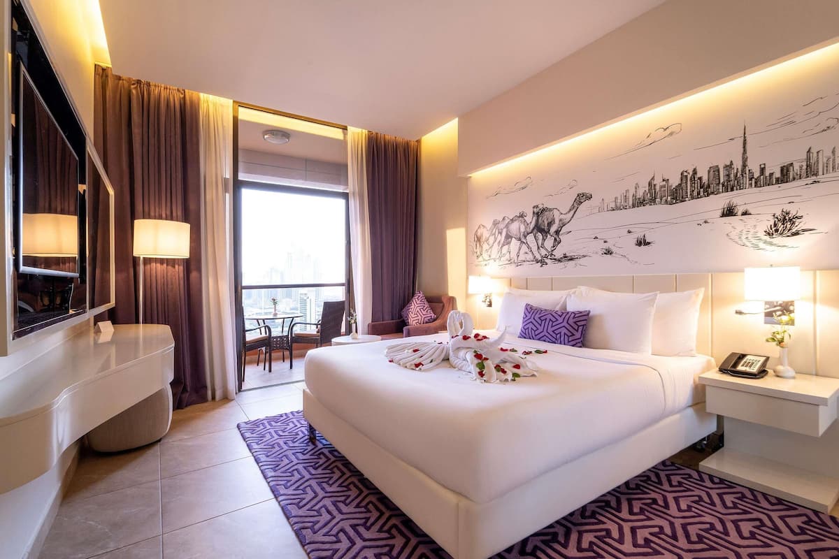A hotel room with a bed, bedside tables, a desk, a chair, a TV, and a cityscape mural.