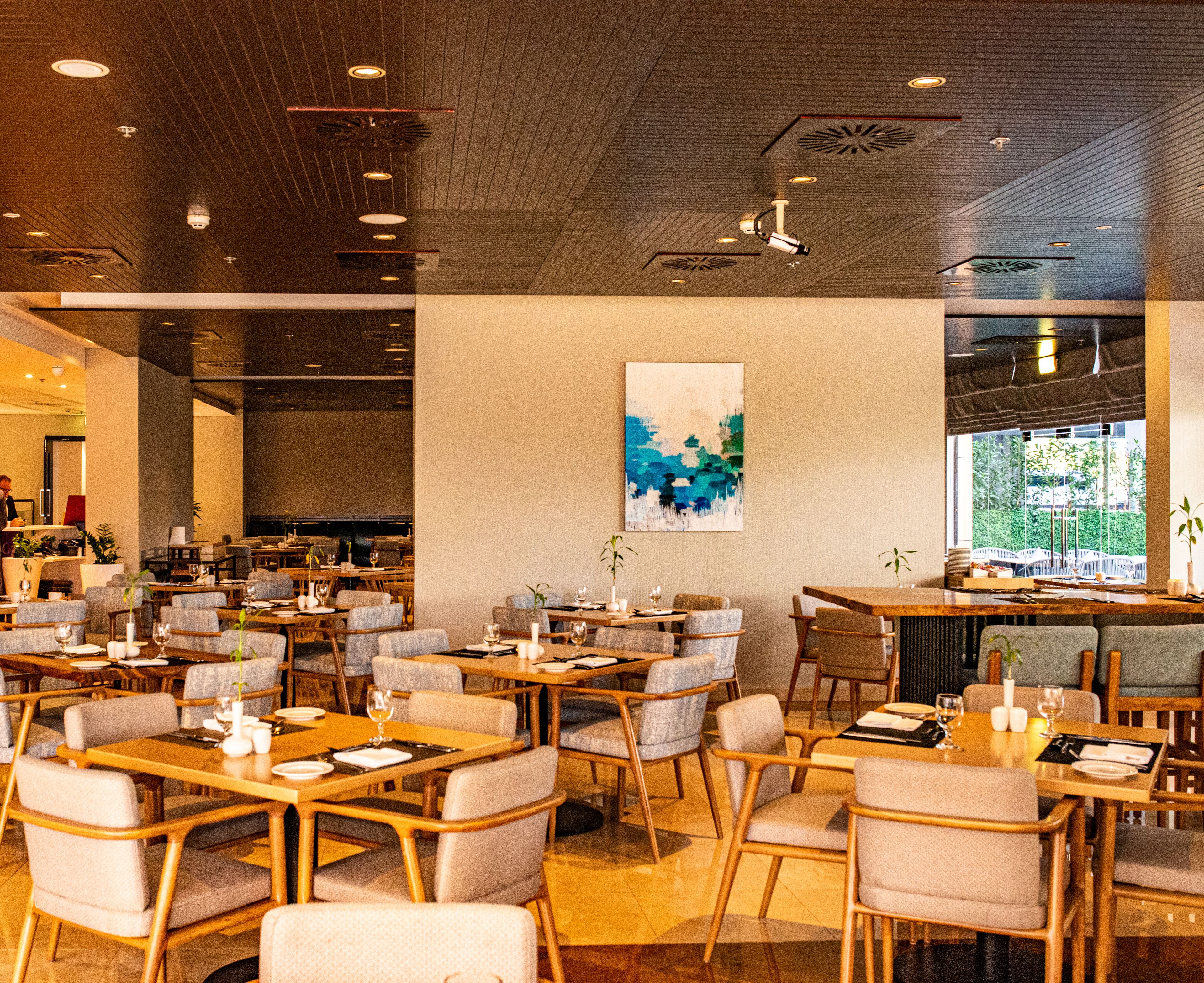 Un restaurante moderno con varias zonas de comedor, mesas y sillas de madera, y un cuadro abstracto grande en la pared.