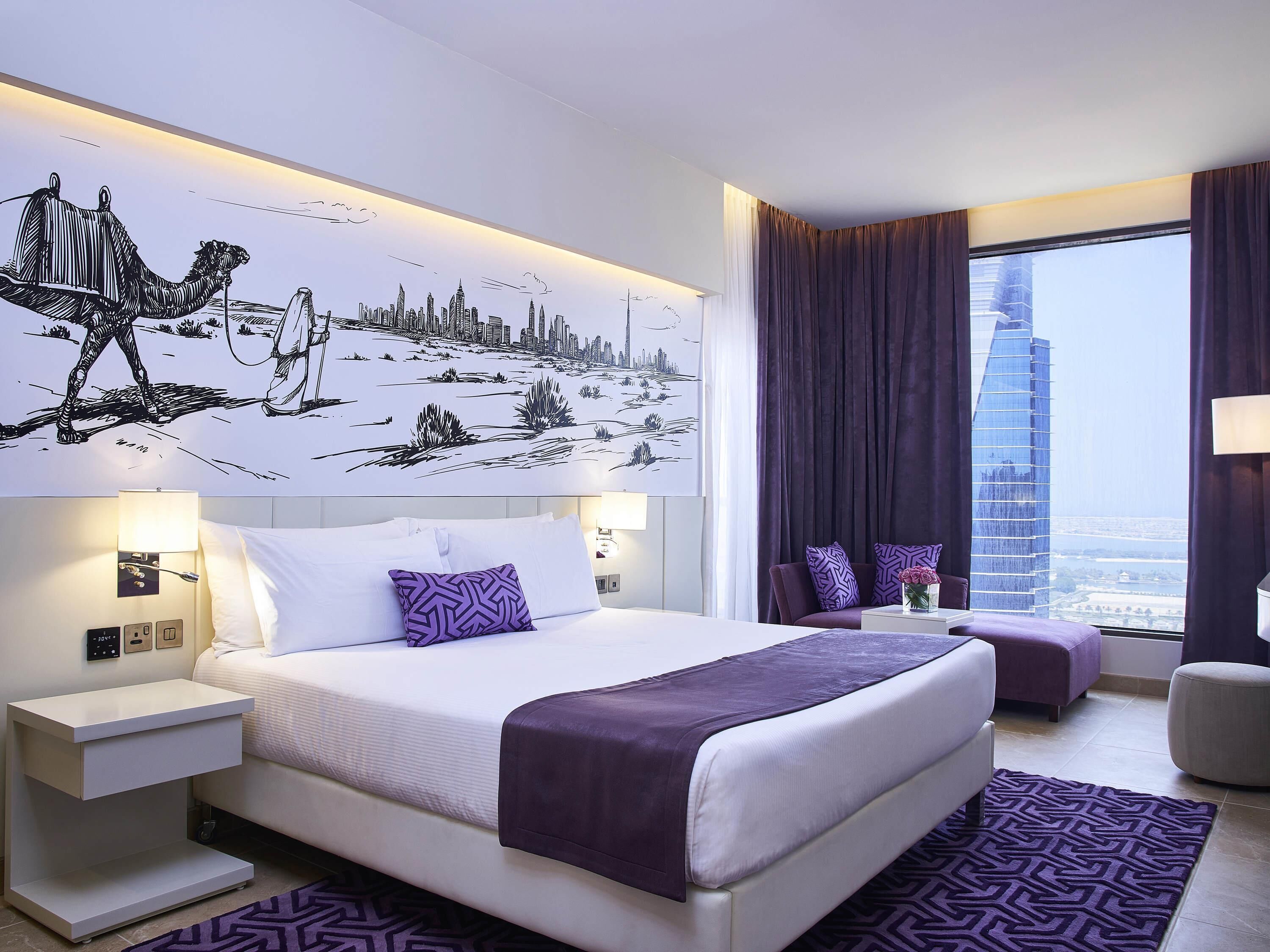 Ein modernes Hotelzimmer mit einem großen Bett, einem Nachttisch, einer Sitzecke mit Blick auf die Stadt und einem Fenster mit Vorhängen.