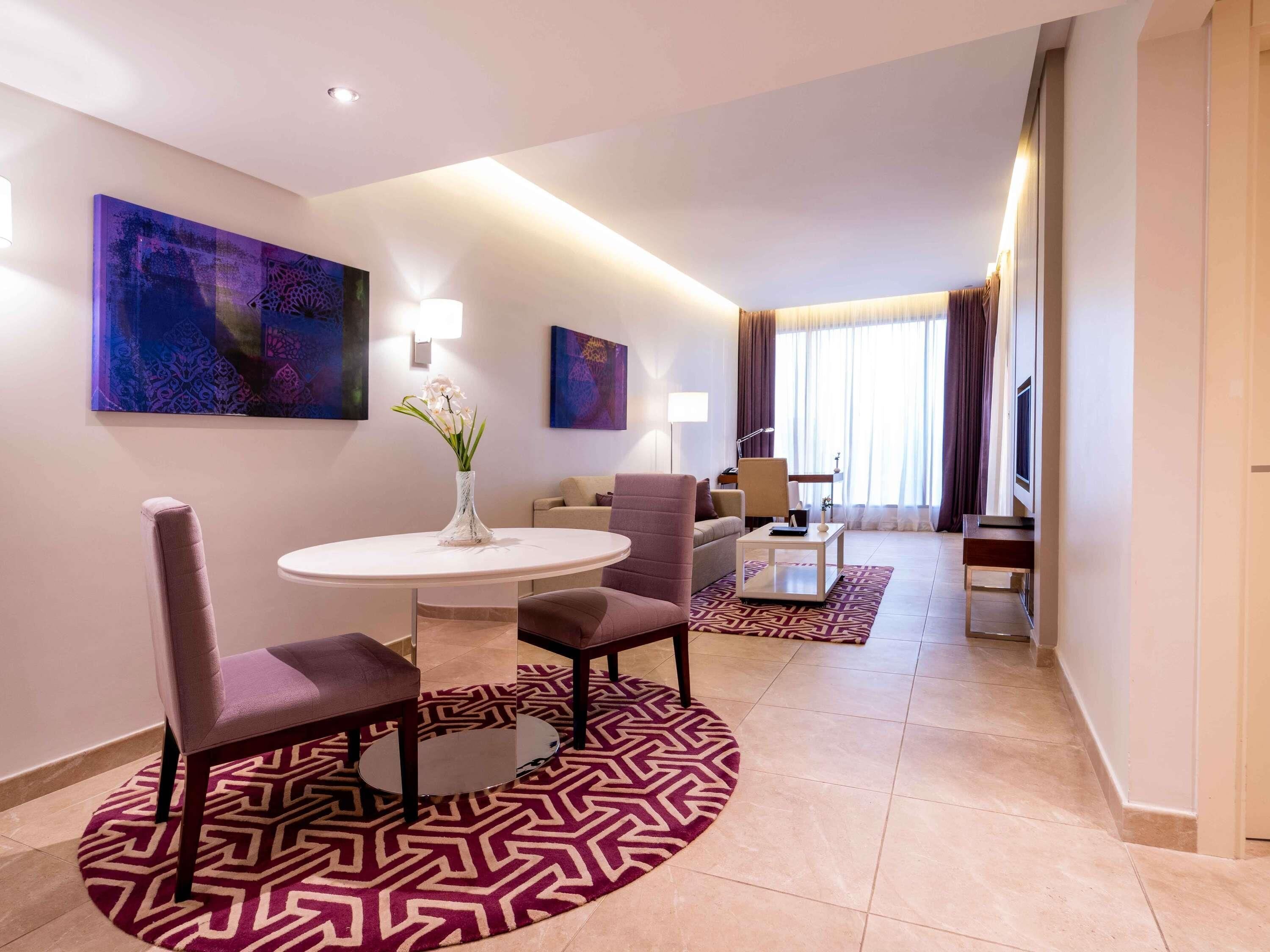 Une chambre d’hôtel moderne avec une table à manger ronde, deux chaises, un vase de fleurs, un canapé, une table basse et des œuvres d’art accrochées aux murs.
