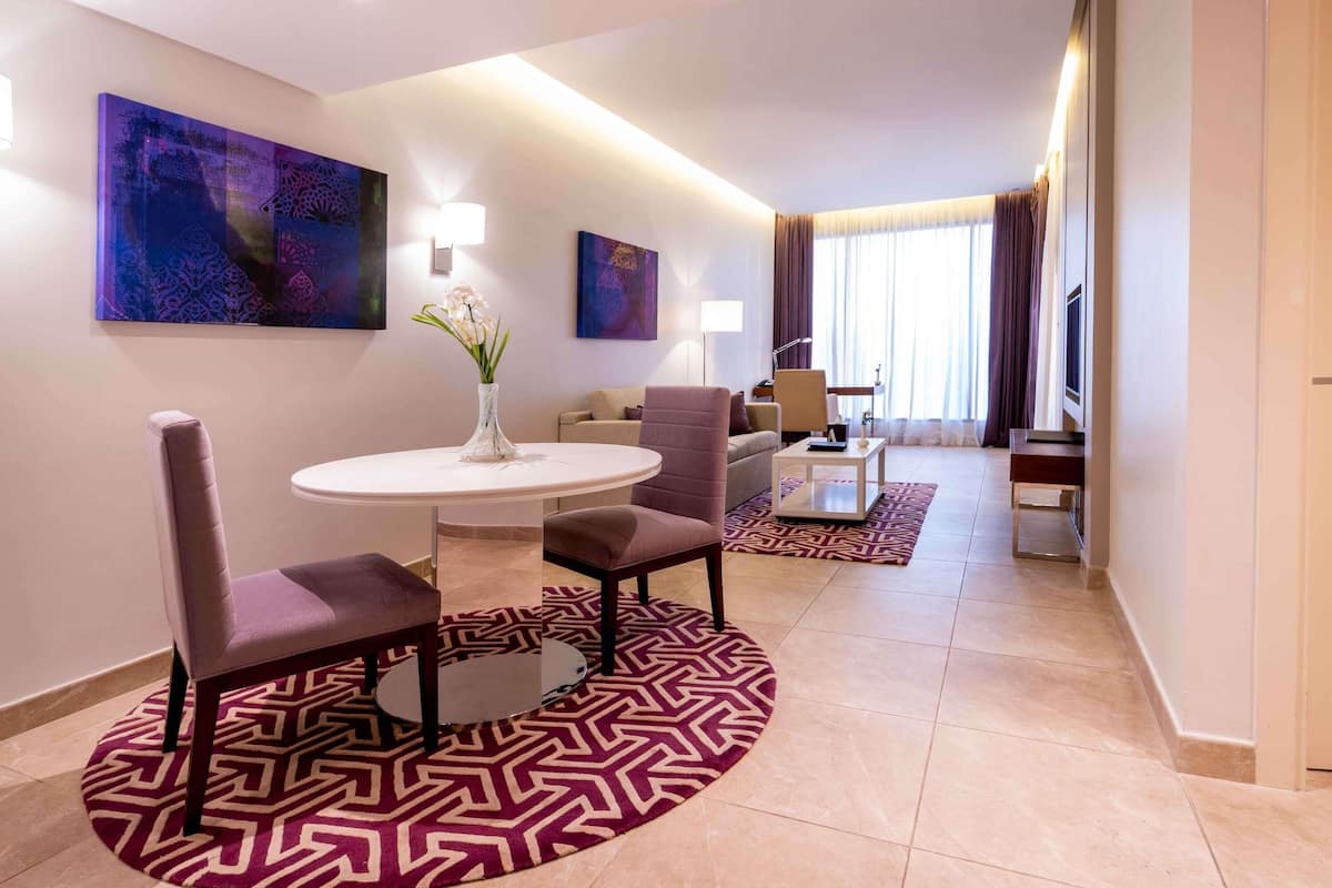 A modern hotel room with a round dining table, two chairs, a vase with flowers, a sofa, a coffee table, and artwork on the walls.