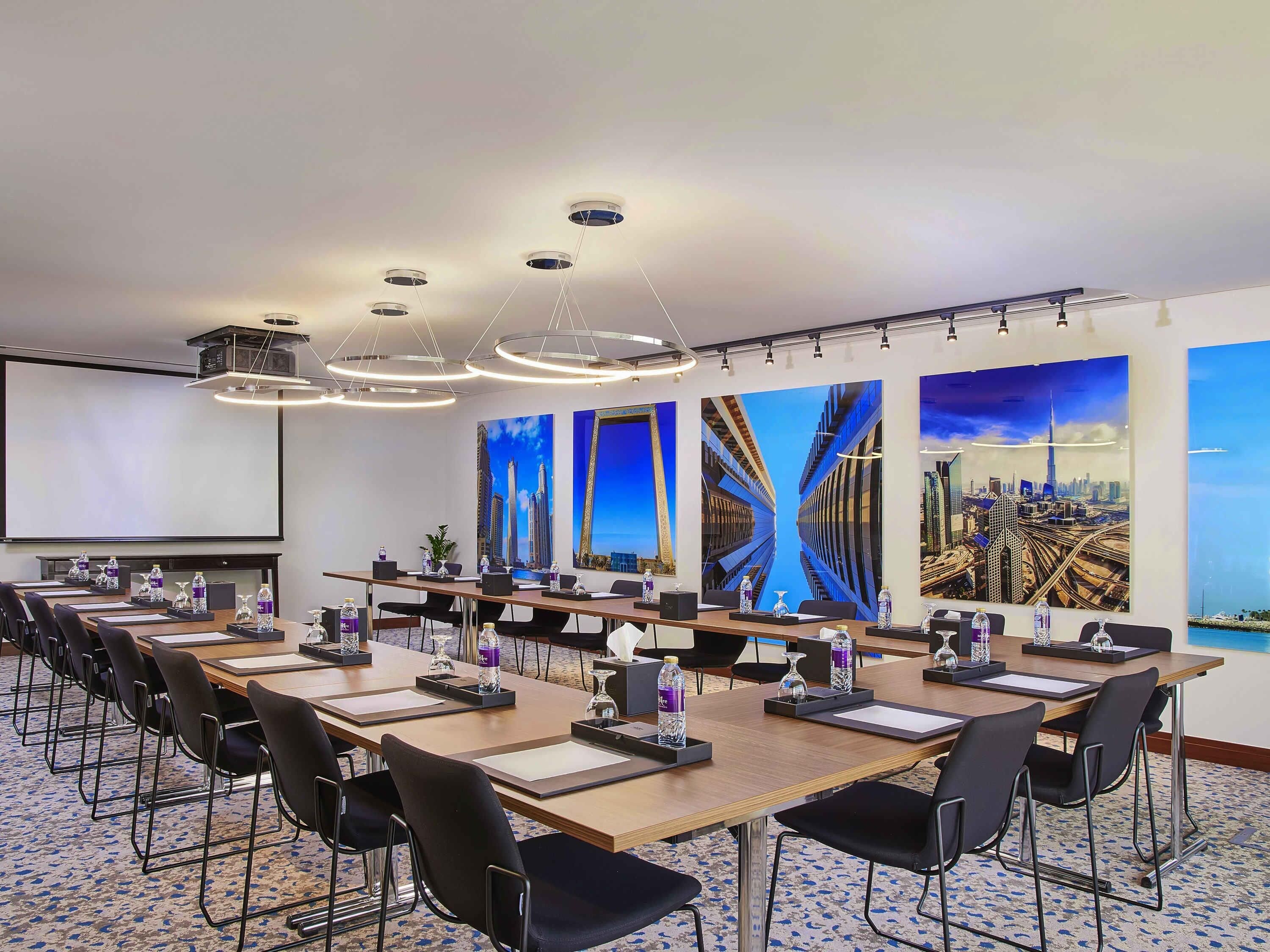 A conference room with multiple tables, chairs, a projector screen, and large cityscape paintings on the walls.