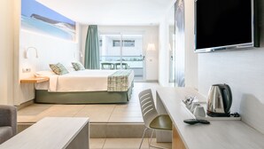 Habitación de hotel con cama, escritorio con hervidor, silla, televisor y balcón con vista al mar.