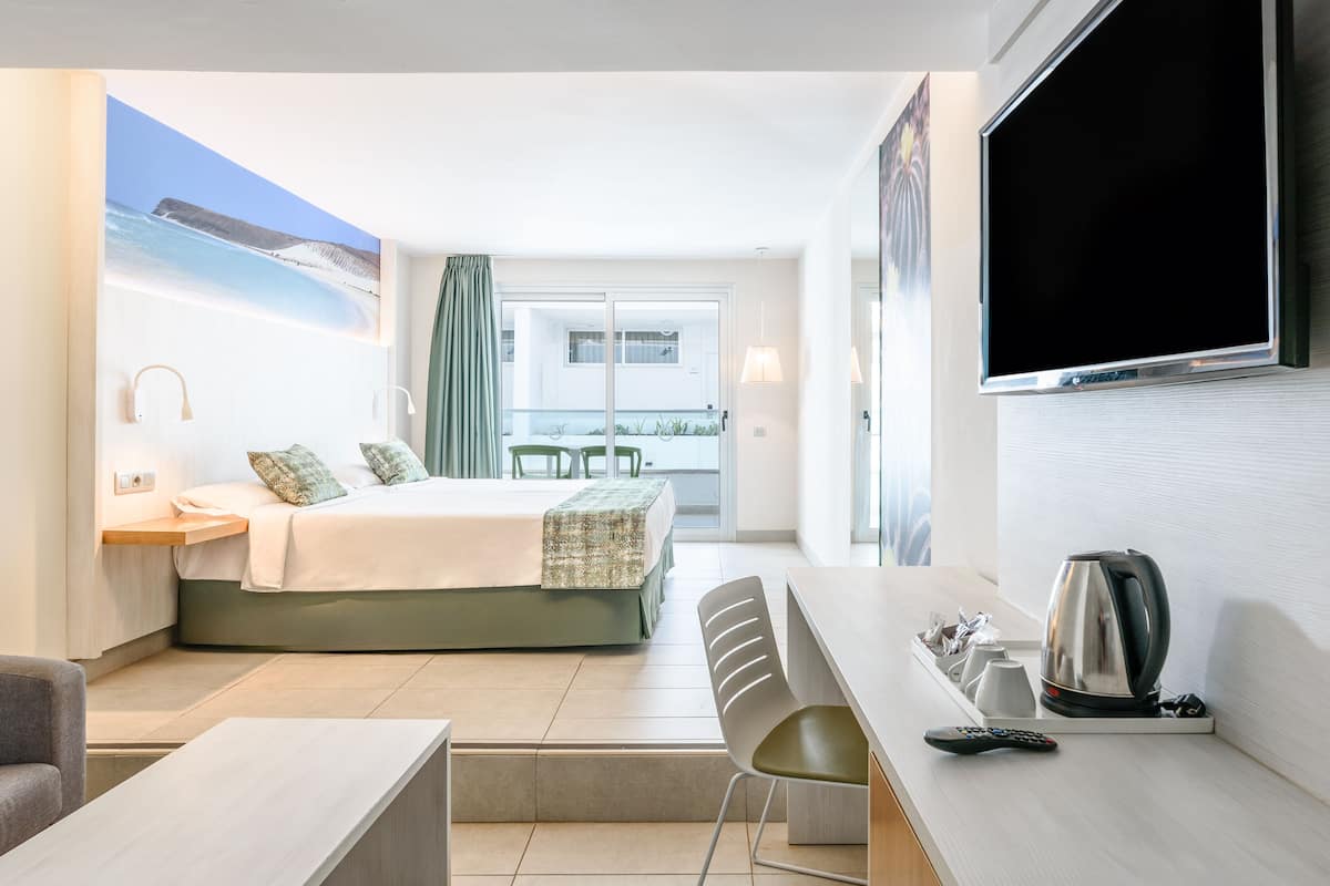 A hotel room with a bed, a desk with a kettle, a chair, a TV, and a balcony with a view of the sea.