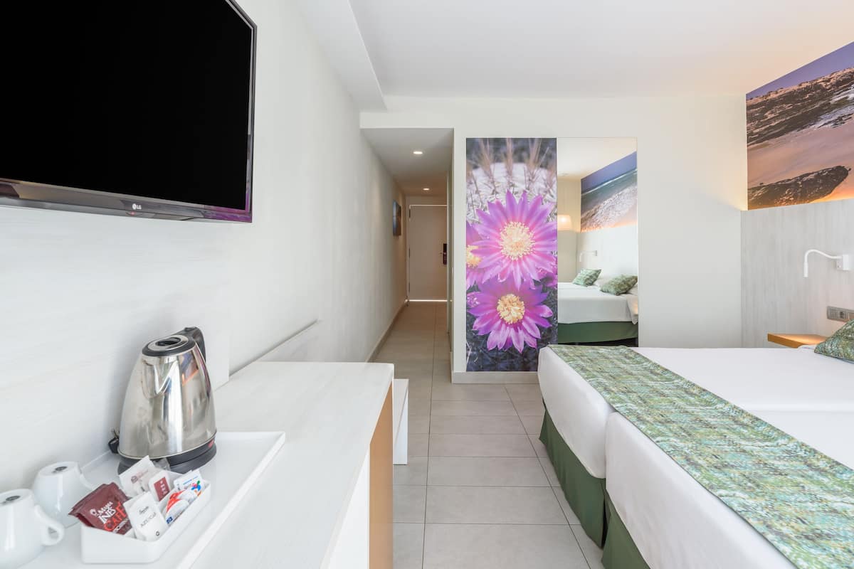 A hotel room with a flat-screen TV, a coffee maker, and a tray with tea bags and a magazine. There is a bed with a green bedspread and a painting of a flower on the wall.
