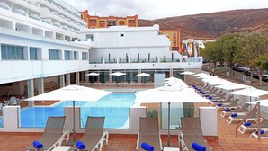 Zona de piscina de hotel con tumbonas y sombrillas, piscina y vistas a una montaña.