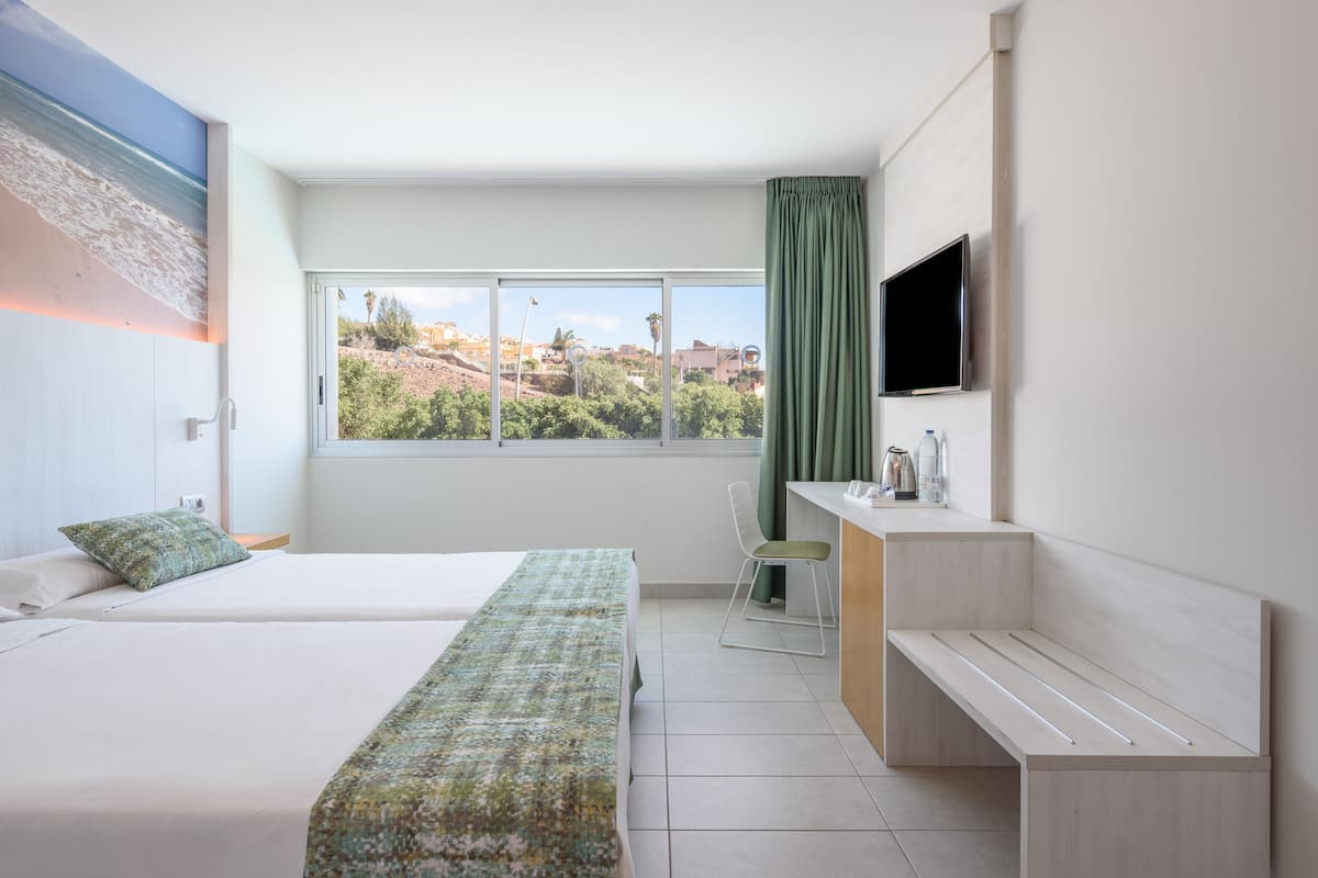 A hotel room with two beds, a desk, a TV, and a window with a city view.