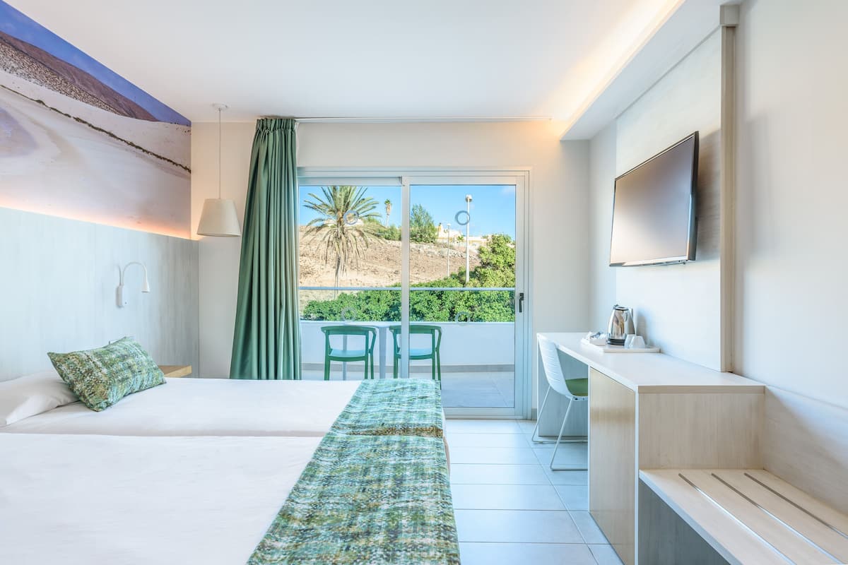 A hotel room with a bed, a desk, a TV, and a balcony with chairs.