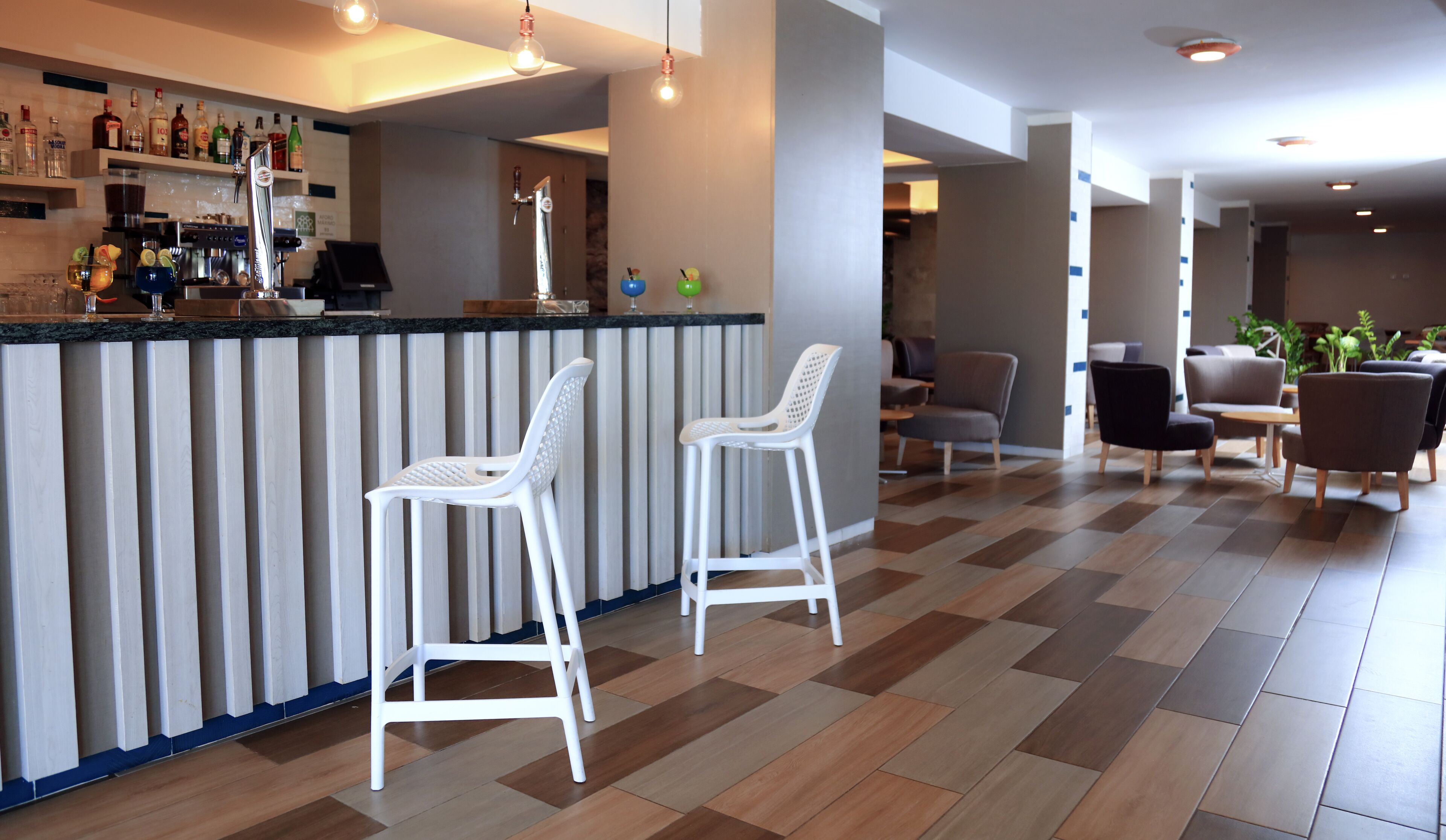 Zona de bar con dos taburetes blancos, una barra con varias botellas y un área de estar con sillas y una mesa.