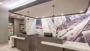 Un mostrador de recepción con una computadora, un refrigerador lleno de bebidas y una pared con un mural texturizado.