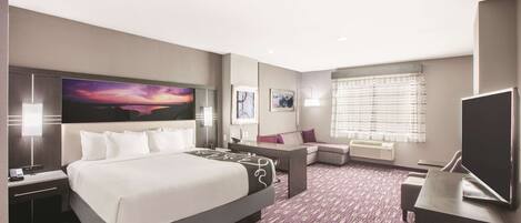 A hotel room with a large bed, a desk, a TV, and a window with blinds.