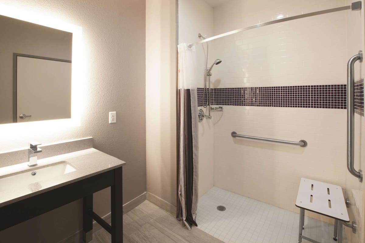 A bathroom with a sink, mirror, shower with grab bars, and a small stool.