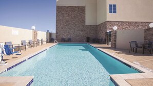 Una piscina rectangular con agua azul cristalina, rodeada de sillas de descanso y mesas, y un muro de piedra con ventanas.