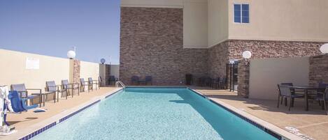 A rectangular pool with clear blue water, surrounded by lounge chairs and tables, and a stone wall with windows.