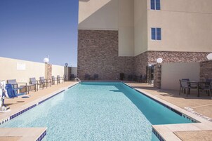 Uma piscina retangular com água azul cristalina, rodeada por espreguiçadeiras e mesas, e uma parede de pedra com janelas.