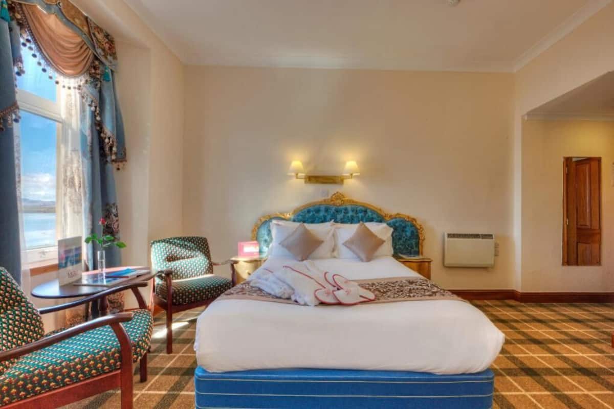 A hotel room with a bed, two chairs, a small table, a window with curtains, and a wall-mounted light fixture.