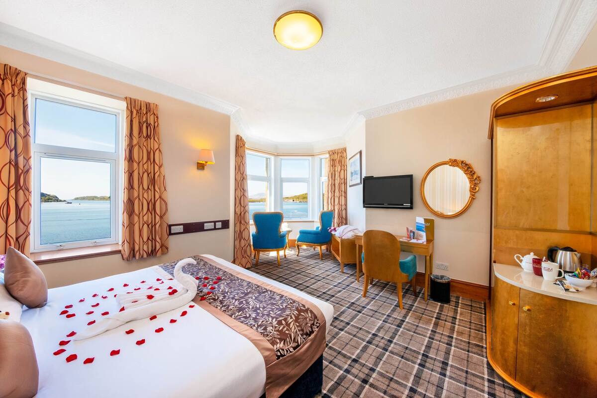 A hotel room with a bed, a desk, a chair, a TV, and a view of the sea.