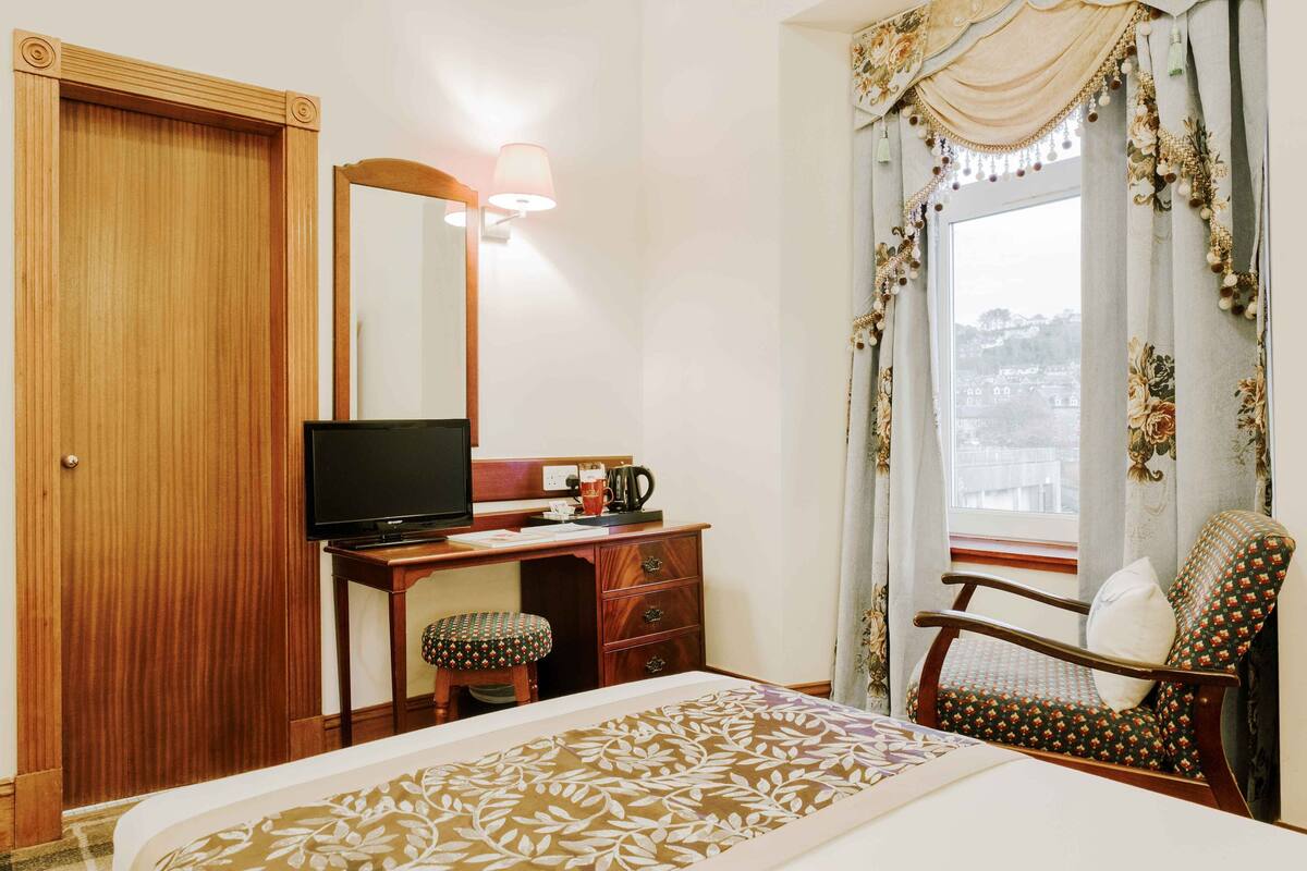 A hotel room with a wooden wardrobe, a desk with a television, a bed with a patterned bedspread, a chair with a cushion, and a window with curtains.