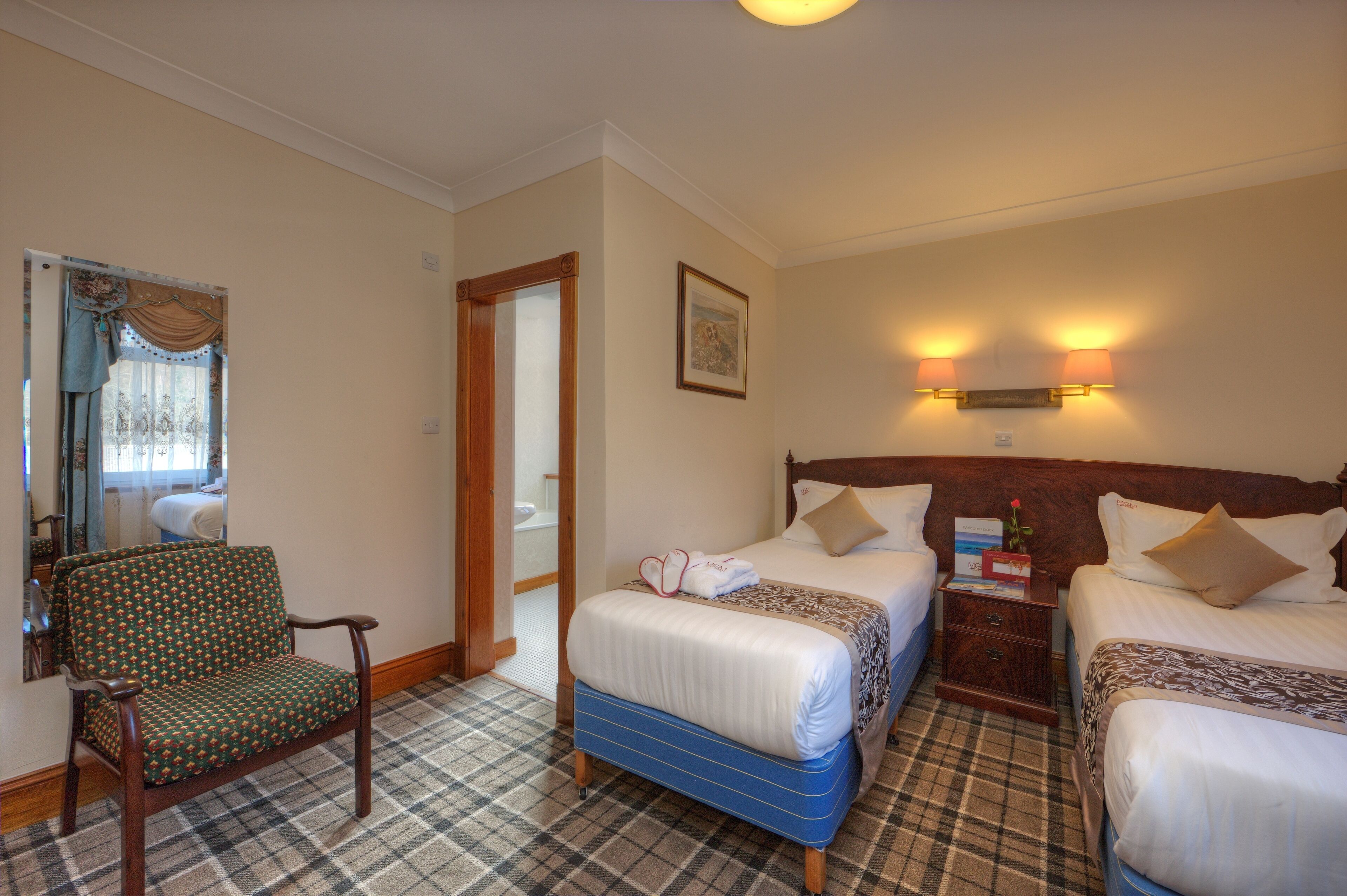 A hotel room with two beds, a chair, a bedside table, a lamp, and a mirror.
