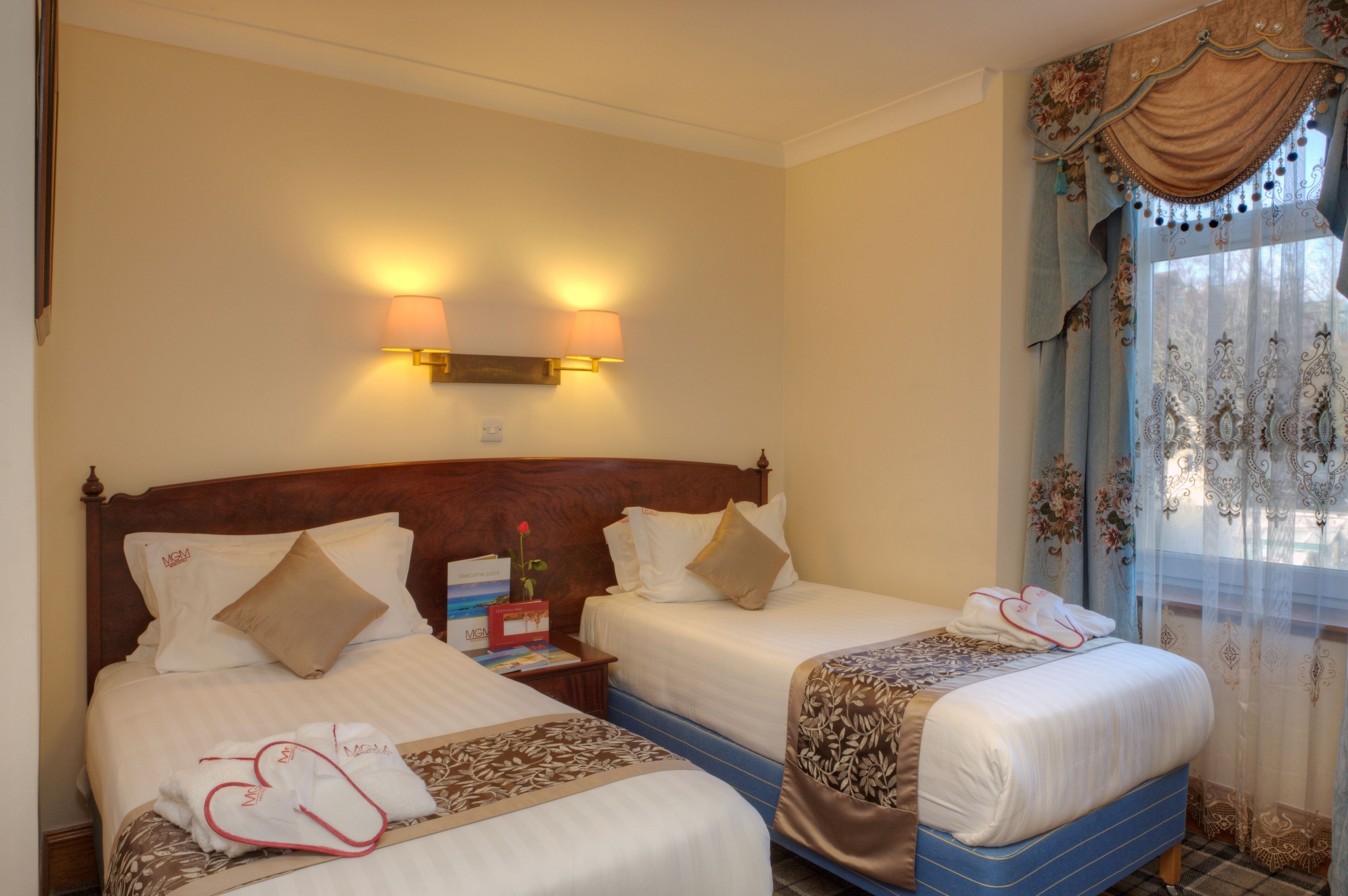 Een hotelkamer met twee bedden, een nachtkastje, een lamp en een raam met gordijnen.
