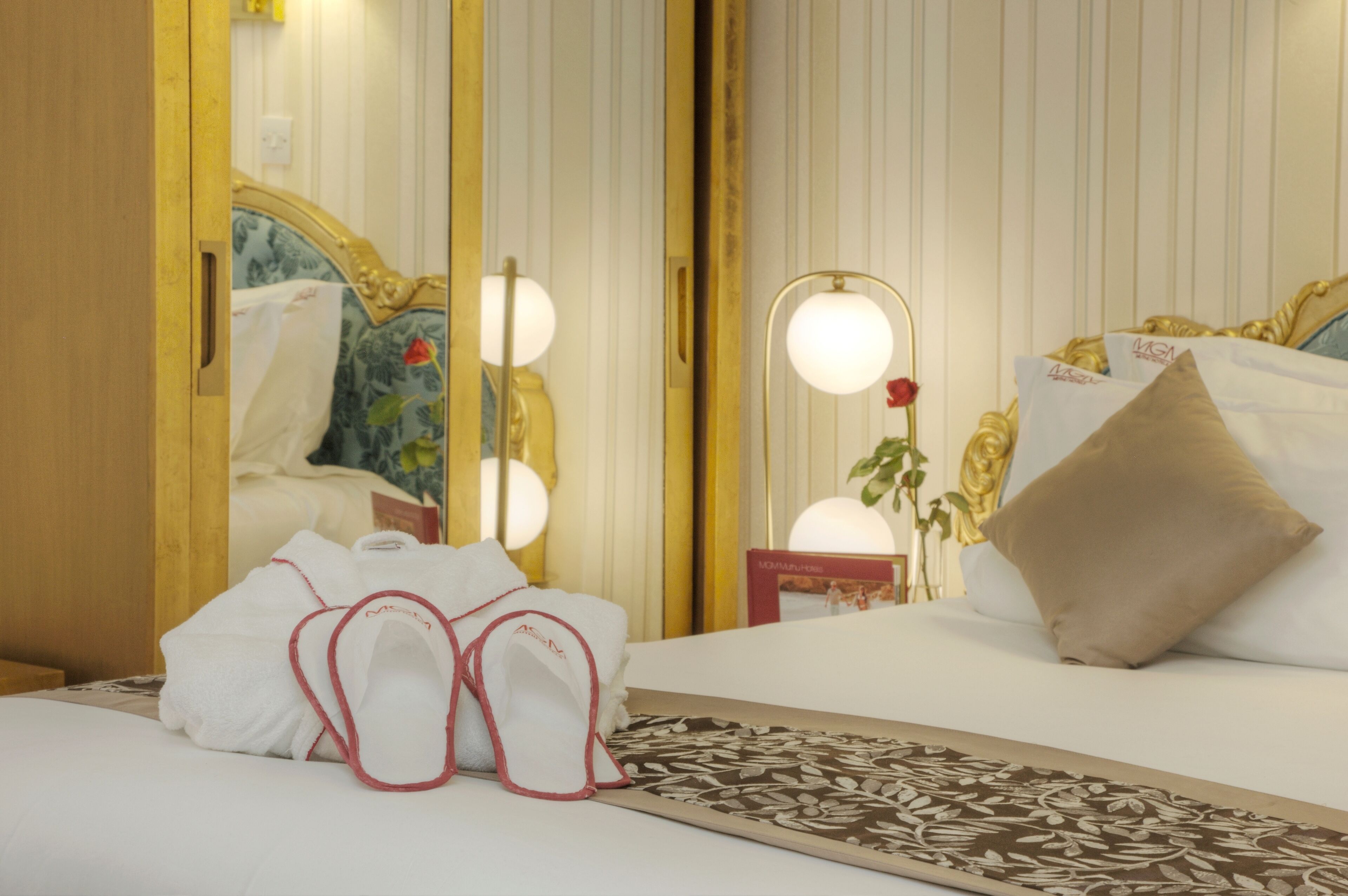 Una camera d'albergo con un letto, uno specchio illuminato, un comodino con un vaso di fiori e un letto rifatto con pantofole.
