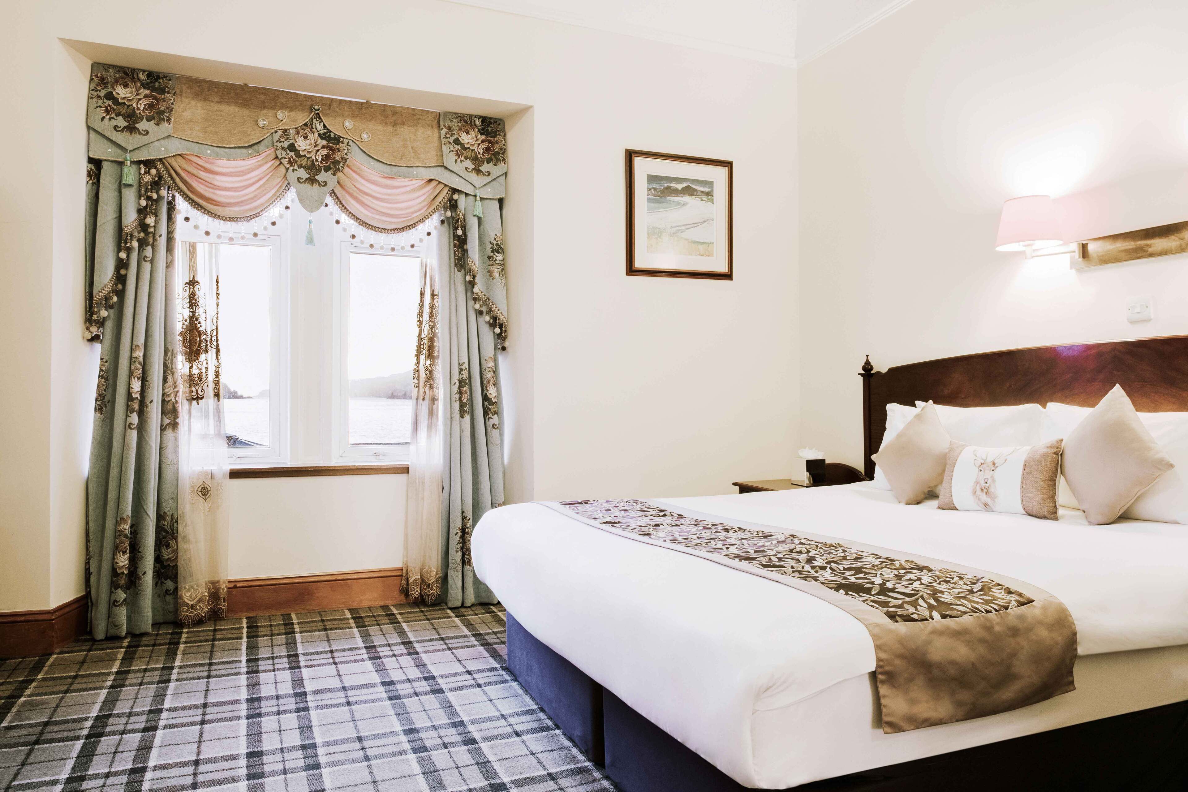 Hotelkamer met een groot bed, een raam met gordijnen, een lijst met een afbeelding aan de muur en een nachtkastje.