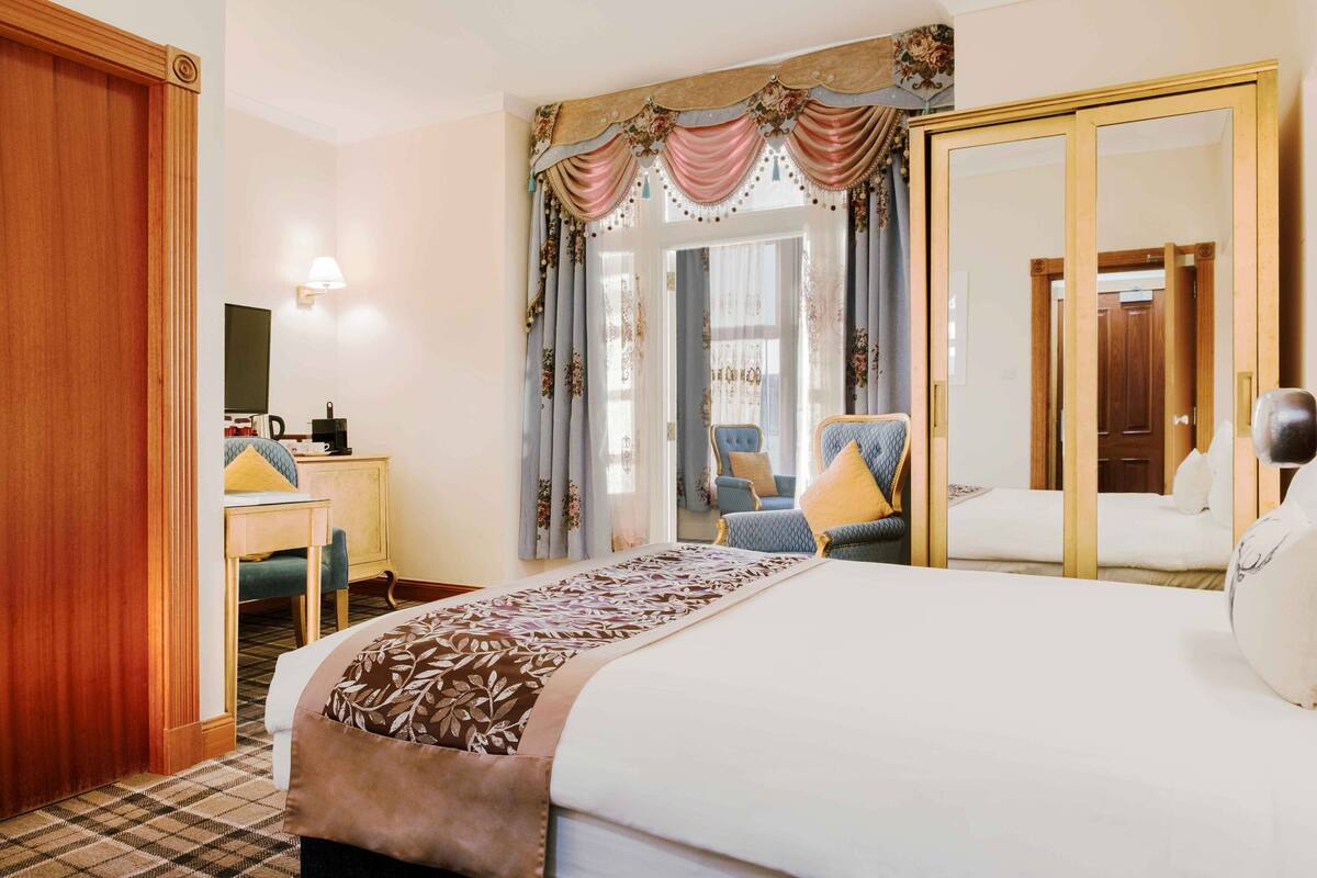 A hotel room with a bed, a desk, a chair, a wardrobe, and a large mirror.