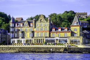 A historic stone building with multiple stories, a prominent entrance with a glass facade, and a flagpole with a flag. - Muthu Alexandra Hotel (Oban)
