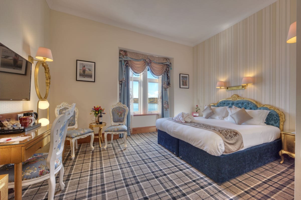 A hotel room with a large bed, a desk with a lamp, a chair, a small table with flowers, and a window with curtains.