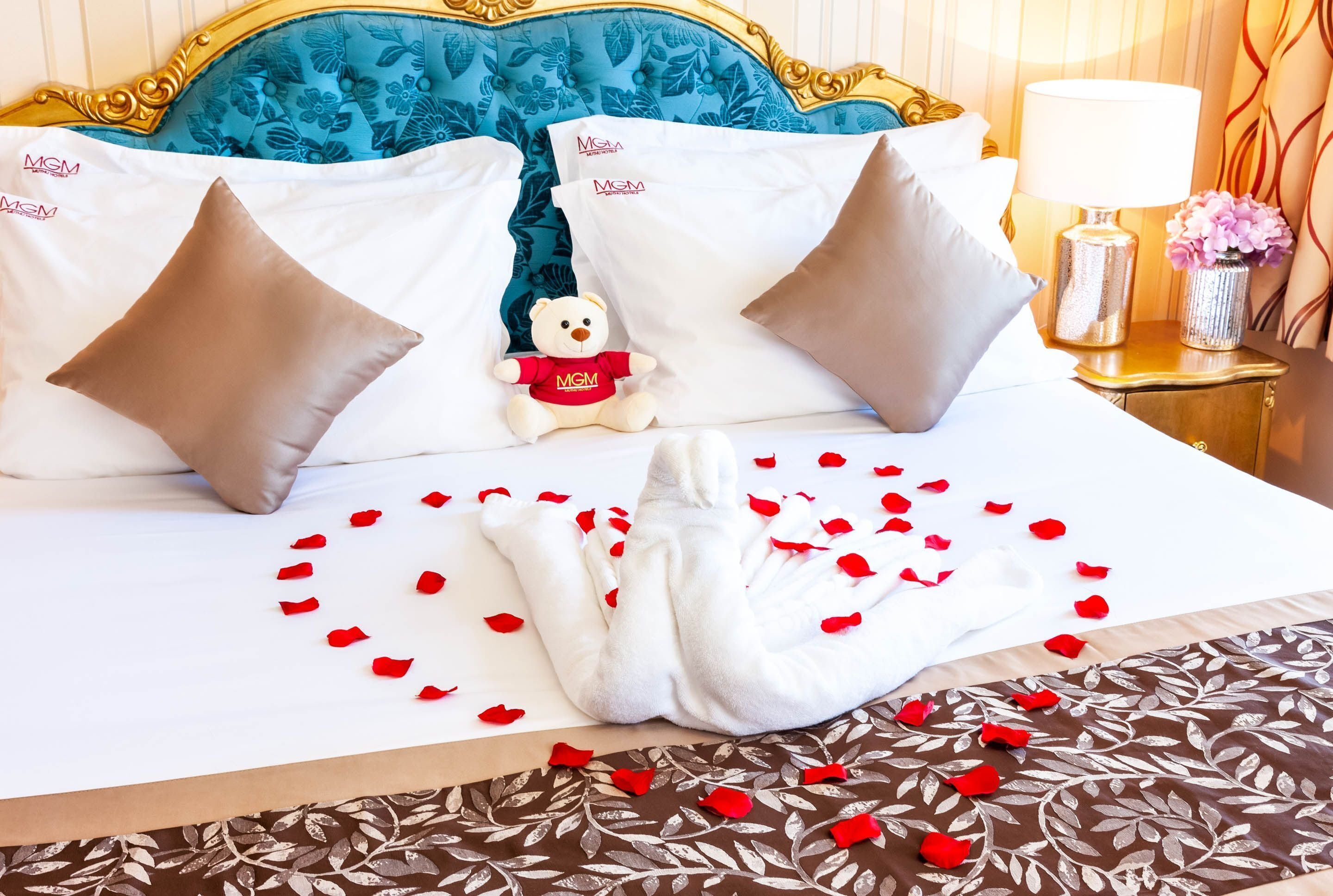 A neatly made bed with white linens, two pillows, a teddy bear, and scattered rose petals.
