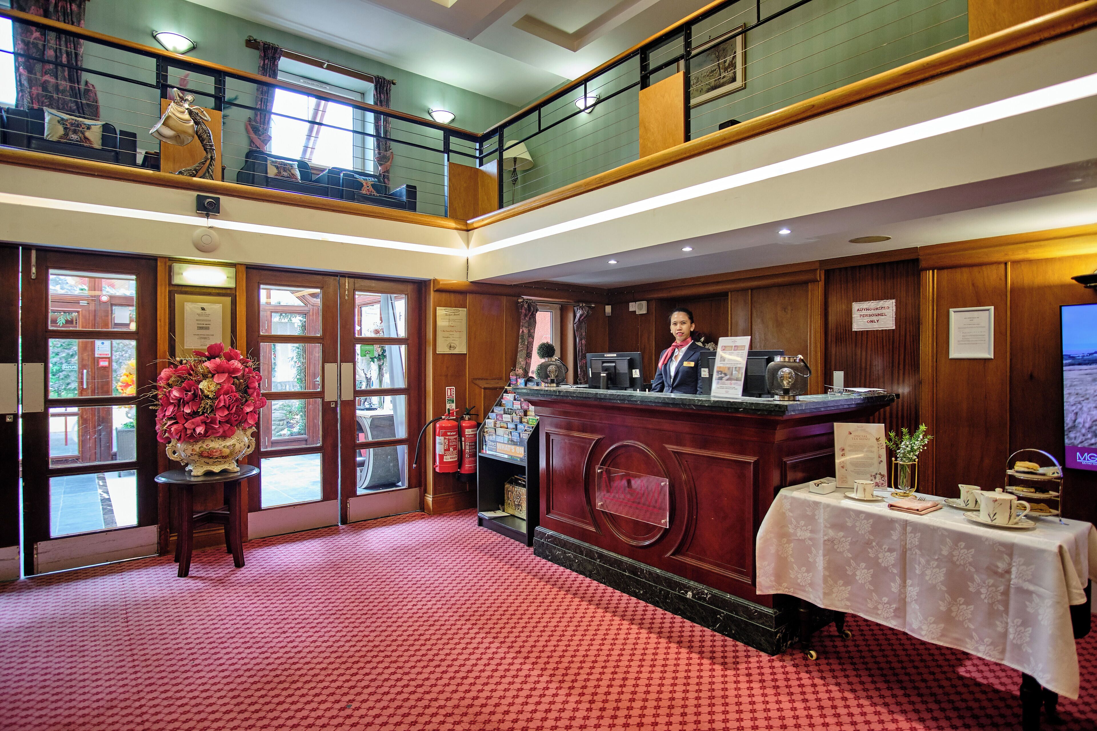 A hotel lobby with a reception desk, a flower arrangement, and a staircase leading to an upper level.