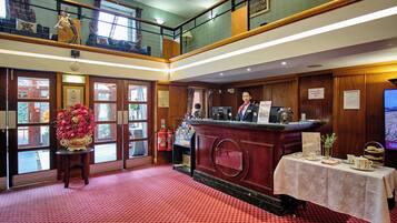 A hotel lobby with a reception desk, a flower arrangement, and a staircase leading to an upper level.