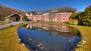 Un gran hotel con su reflejo en un estanque, rodeado de vegetación y con una montaña al fondo.