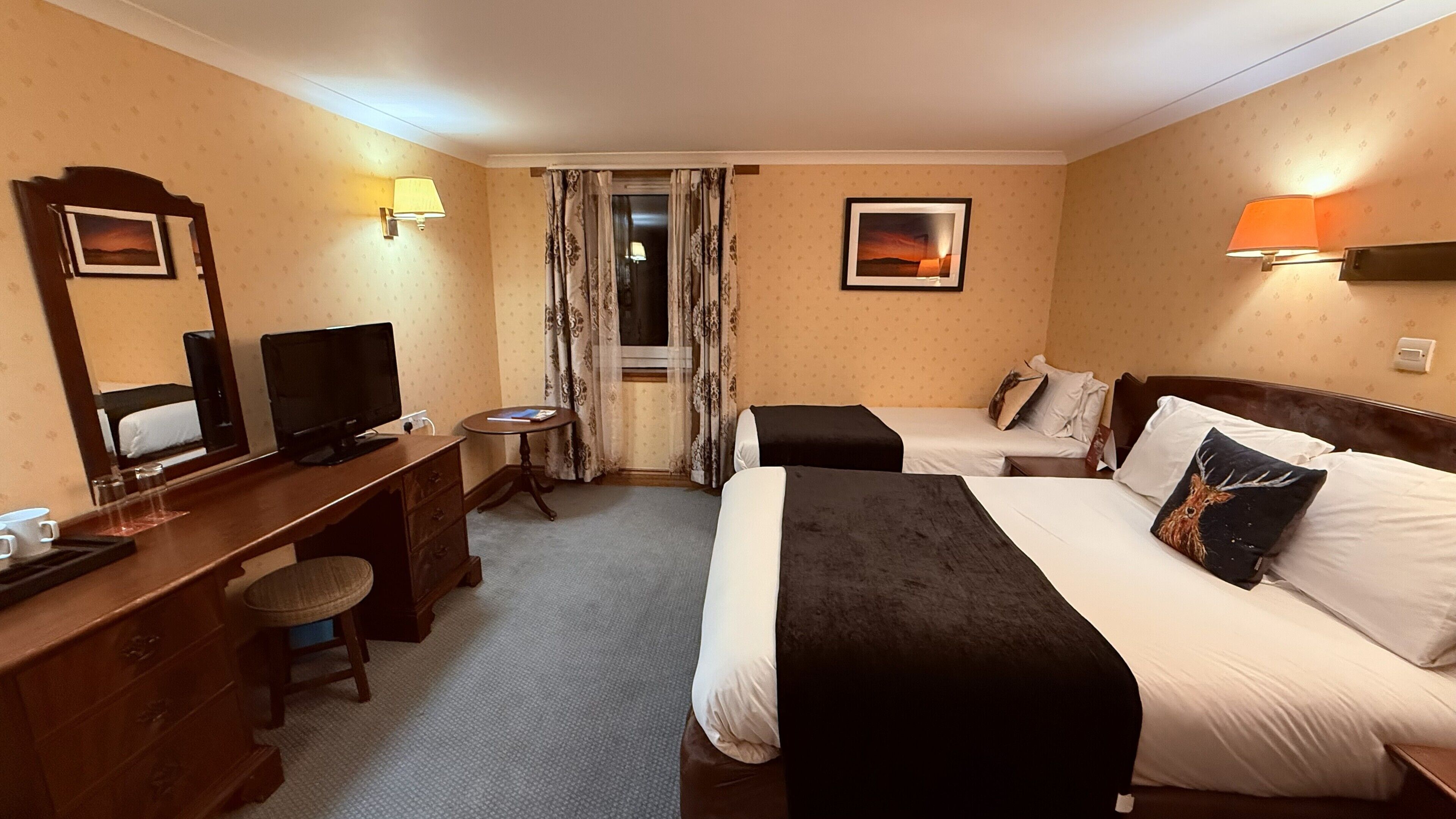 A hotel room with two beds, a desk with a TV, a small table, and a window with curtains.