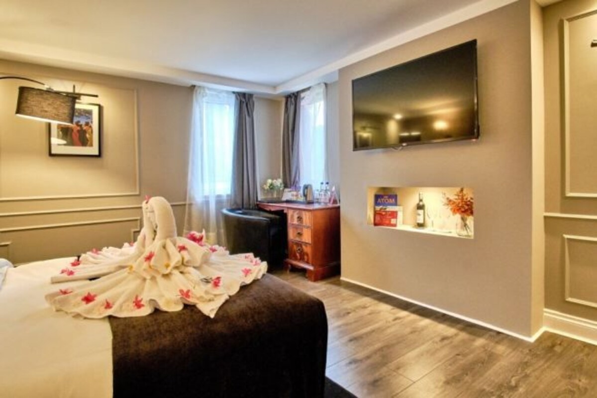 A hotel room with a bed, a desk, a flat-screen TV, a framed picture on the wall, and a small shelf with items.