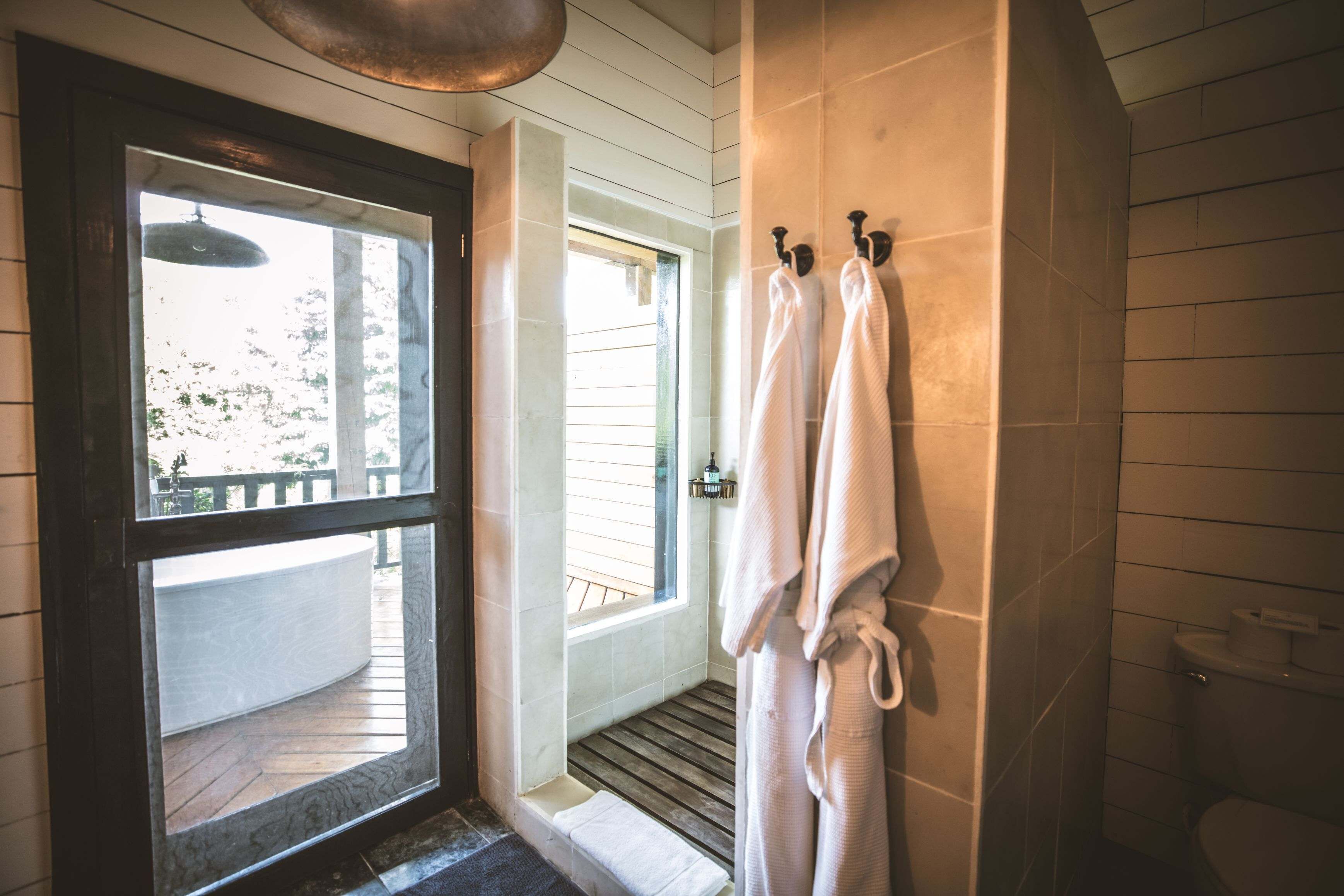 Baño con suelo de madera, zona de ducha y inodoro. Hay toallas colgadas en ganchos y una ventana que proporciona luz natural.