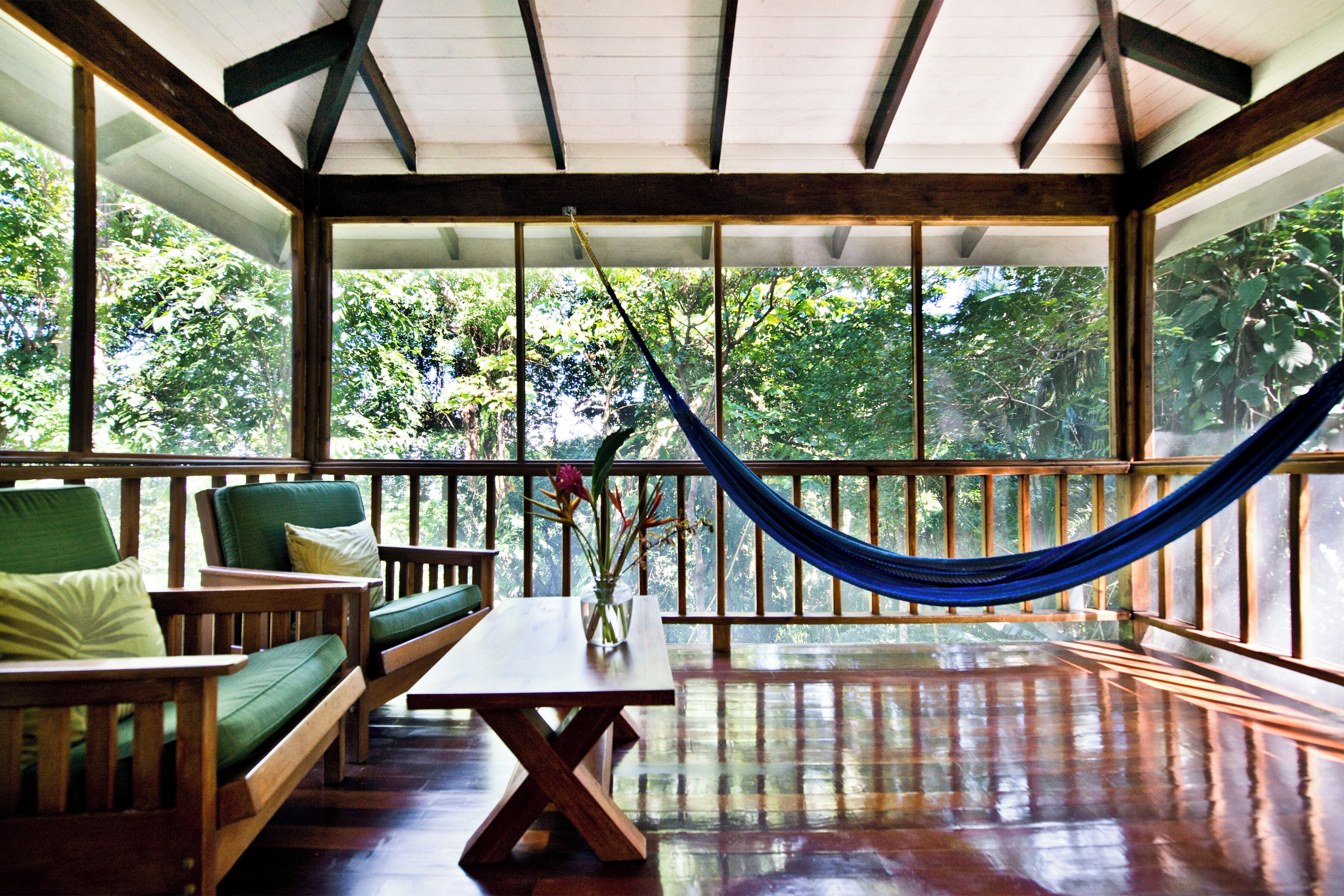 A wooden deck with a hammock, cushioned seating, and a table with a vase of flowers.