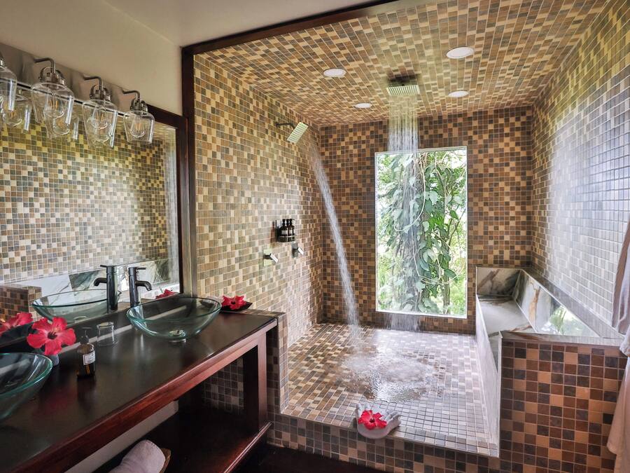 A bathroom with a glass bowl sink, a shower with a tiled wall, and a window with a view of greenery.