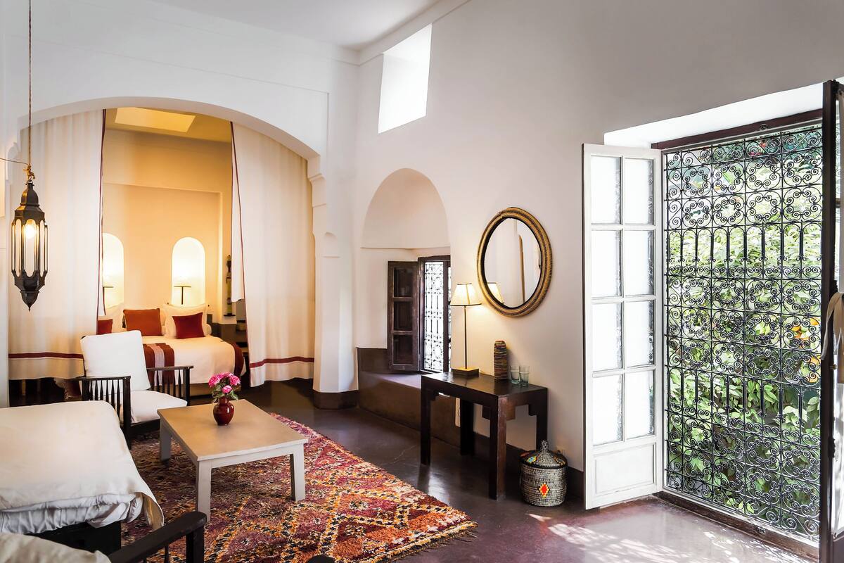 A hotel room with a bed, a sitting area with a table and chairs, a small table with a vase of flowers, a mirror on the wall, and a large window with a decorative iron gate.