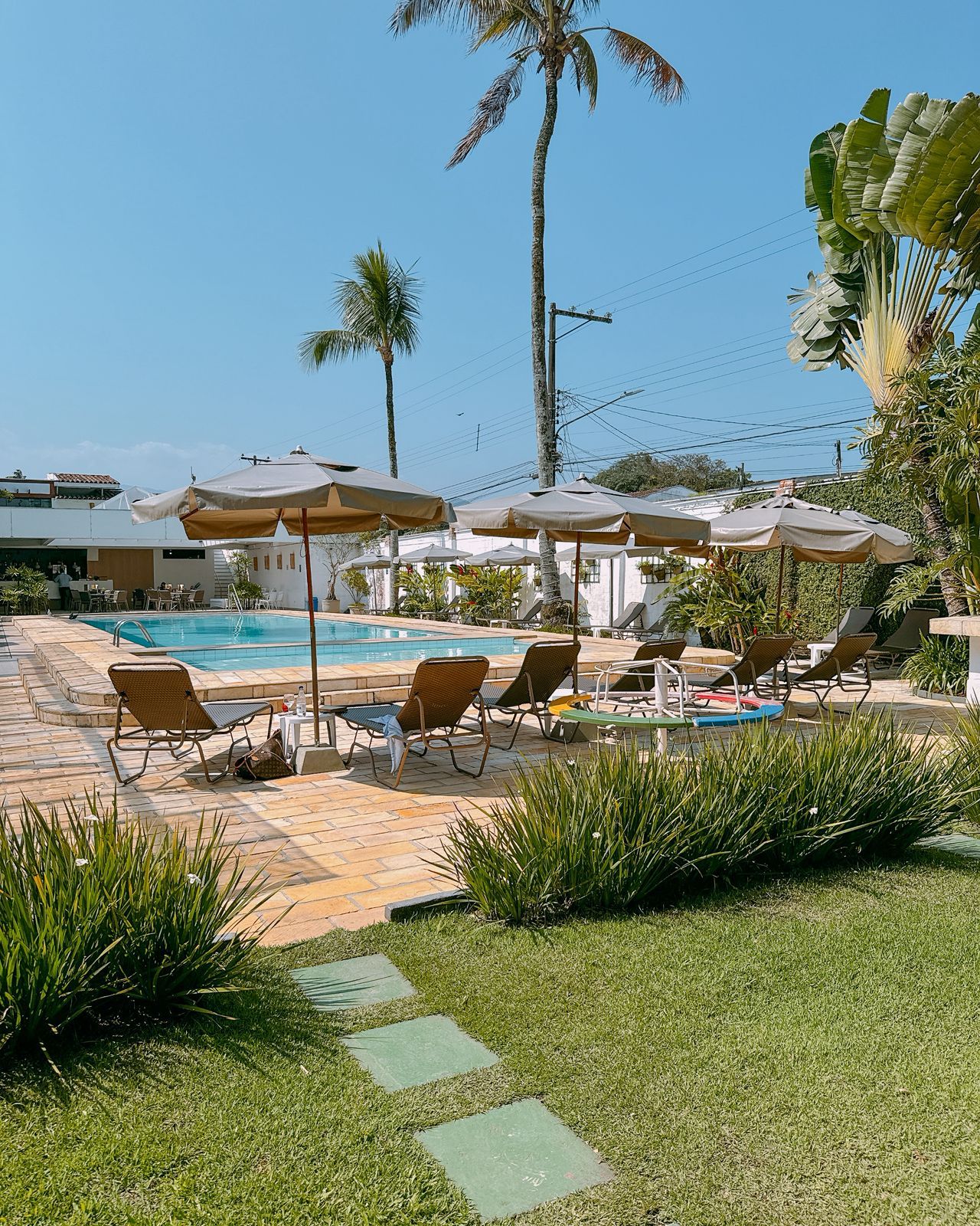 Área ao lado da piscina com espreguiçadeiras e guarda-sóis, rodeada por palmeiras e vegetação.
