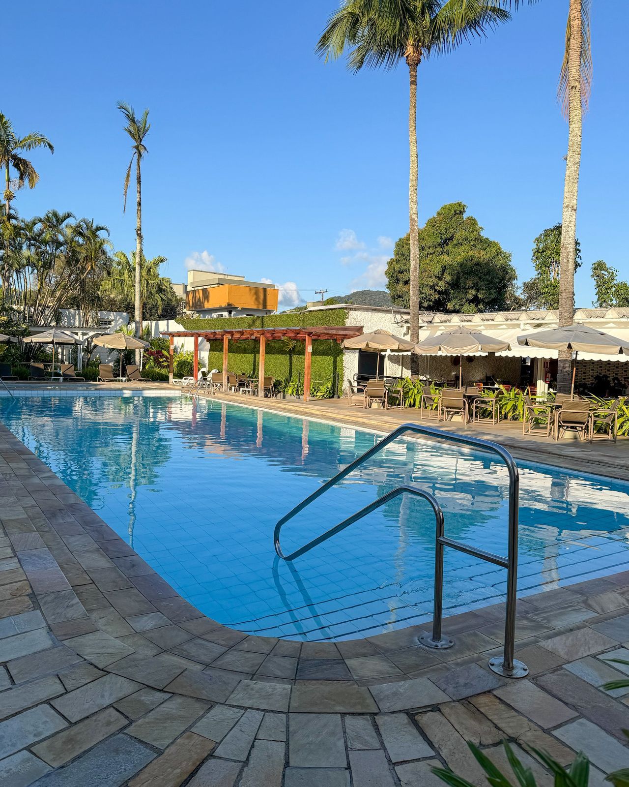 A swimming pool with a tiled deck, surrounded by palm trees and a building with outdoor seating.