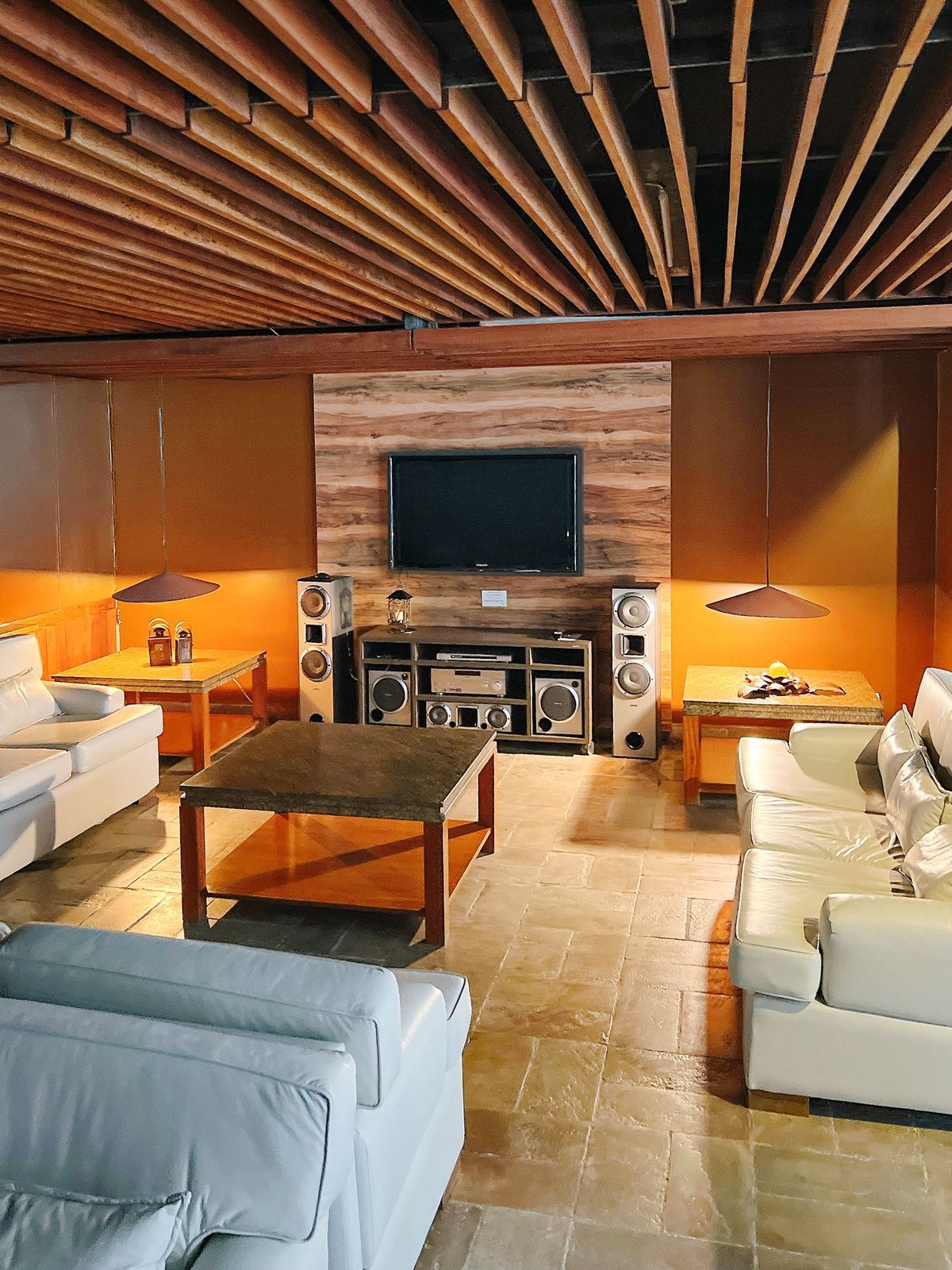 A modern living room with a flat-screen TV mounted on a wooden wall, a coffee table, two white sofas, and a stone-tiled floor.