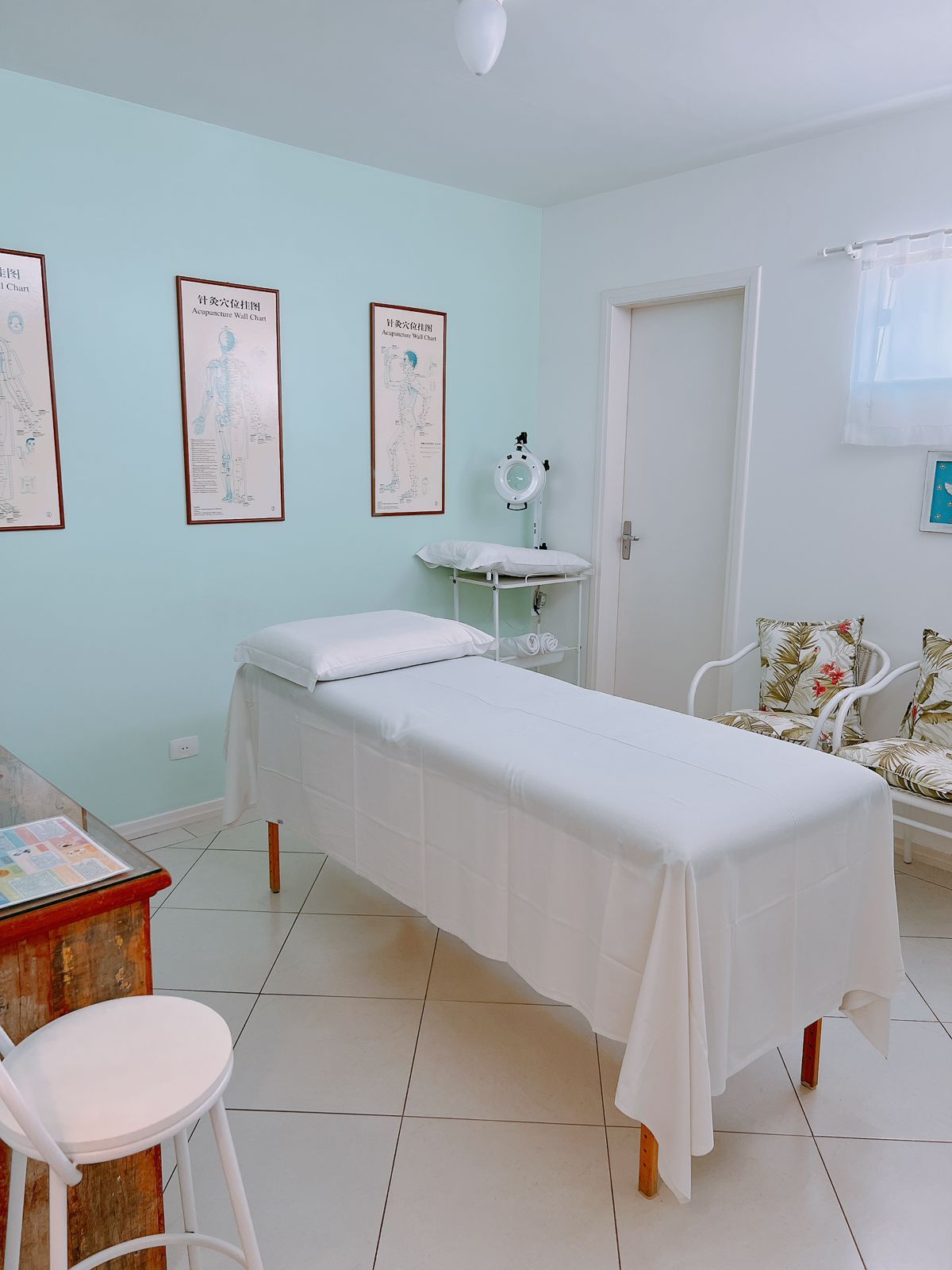 Une salle de consultation équipée d’un lit, d’une petite table et d’une chaise. Des affiches sont accrochées au mur, et une porte se trouve sur le côté droit.