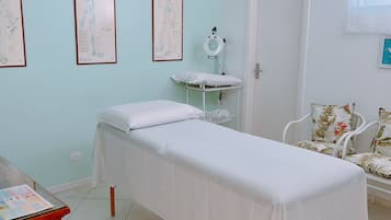 Une salle de consultation équipée d’un lit, d’une petite table et d’une chaise. Des affiches sont accrochées au mur, et une porte se trouve sur le côté droit.
