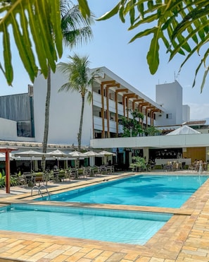 Una piscina con solárium empedrado, rodeada de mobiliario exterior y sombrillas, con un edificio al fondo.