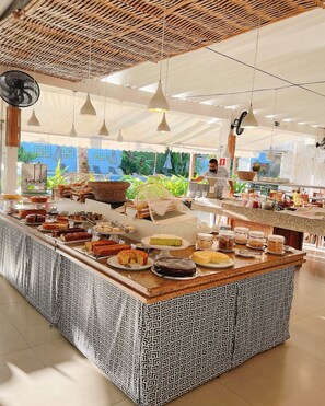 A buffet with various food items, a patterned tablecloth, hanging lights, and a view of the outdoors. - Ubatuba Palace Hotel (Ubatuba)